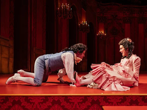A man in 18th-century costume kneels on the floor, holding the ankle of a seated woman in a pink period dress, on a red-lit stage with ornate decor and chandeliers.