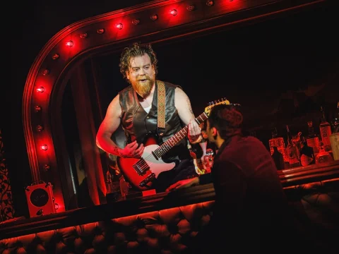 A man with a beard plays an electric guitar on a bar counter, while another person in a dark jacket leans against the bar listening, under a red-lit marquee.