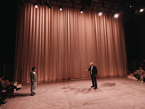 Two people stand facing each other on a stage with wooden flooring and a large beige curtain, while several audience members watch from the sides.