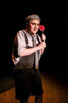 A person in period costume holds a pink flower on stage under dramatic lighting.