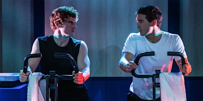 Two men are riding stationary bikes indoors, facing each other with towels draped over the handlebars.