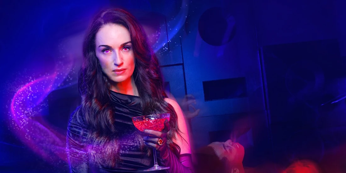 A woman with long dark hair in a black dress holds a red drink, standing in a dimly lit, blue and purple-toned room.