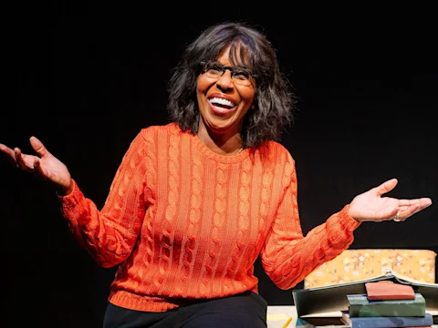 A woman wearing glasses and an orange sweater sits smiling with arms raised, surrounded by books on a table, against a dark background.