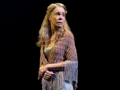 A woman with long, wavy blonde hair stands on stage wearing a fringed shawl, looking off to the side with a serious expression, against a black background.