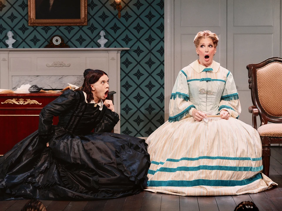 Two actors in period costumes on stage. One, in a black dress, crouches with a shock expression, while the other, in a white and teal dress, sits with an equally surprised expression.