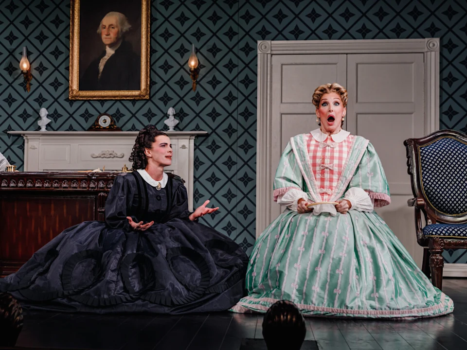 Two actors in period costumes on stage. One, in a black dress, crouches with a shock expression, while the other, in a white and teal dress, sits with an equally surprised expression.