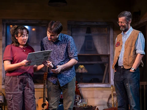Three people stand indoors; a woman holds an open book while two men look at it. The background includes a guitar, lamp, and window with curtains.