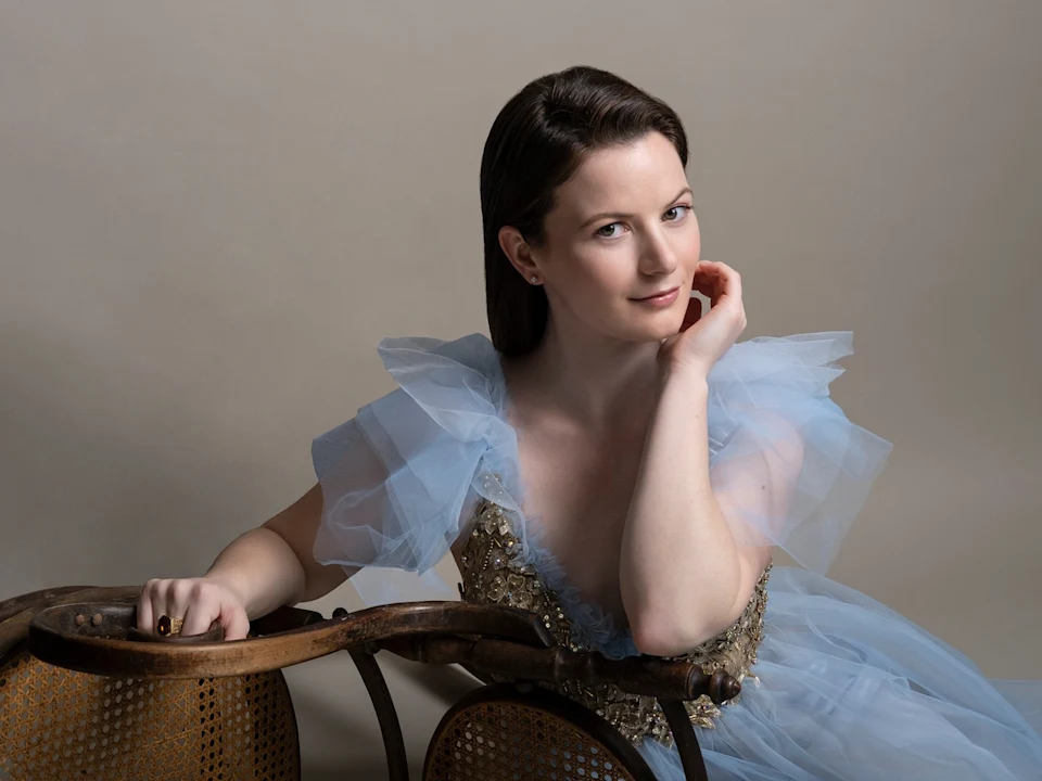 A woman in a blue tulle dress with gold embellishments sits behind a wooden chair, resting her chin on her hand and looking at the camera against a plain background.
