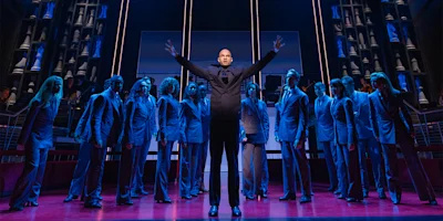 A man stands center stage with arms raised, surrounded by people in suits on a lit theater stage with dramatic lighting and set design.