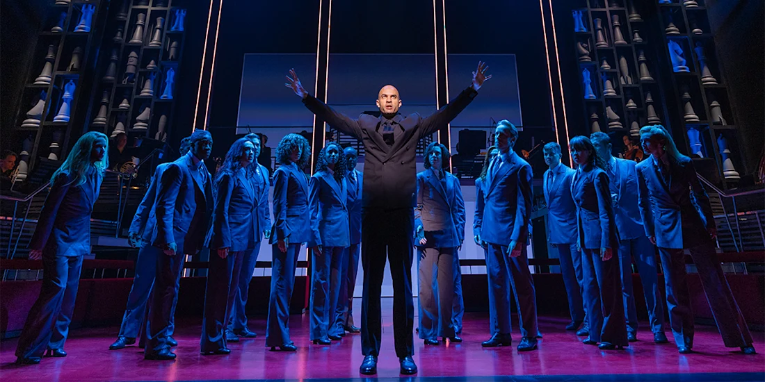 A man stands center stage with arms raised, surrounded by people in suits on a lit theater stage with dramatic lighting and set design.