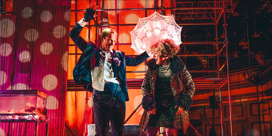 Two people in formal and eccentric clothing stand on a vibrant stage; one holds a lacy umbrella over the other’s head. The background features scaffolding and dramatic lighting.