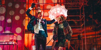 Two people in formal and eccentric clothing stand on a vibrant stage; one holds a lacy umbrella over the other’s head. The background features scaffolding and dramatic lighting.