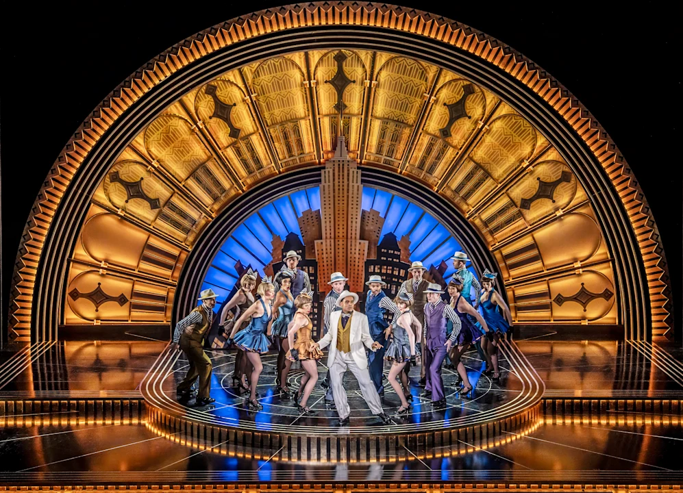 A group of performers in colorful costumes pose in front of a gold and blue Art Deco stage set featuring a cityscape and dramatic lighting.