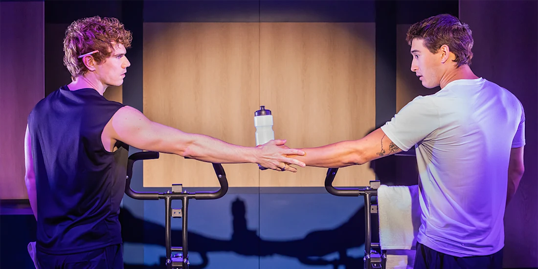 Two men standing beside stationary bikes, one handing a water bottle to the other. Both are dressed in athletic wear and facing each other.