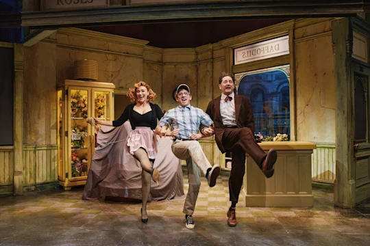 Three actors perform a choreographed kick in a vintage flower shop stage set, smiling and holding hands mid-step.