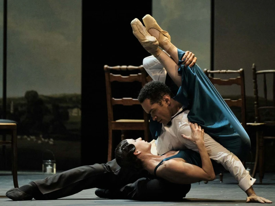 Two ballet dancers perform an intricate lift on stage. The male dancer supports the female dancer who is upside-down, her legs over his shoulder, both maintaining a poised and focused expression.