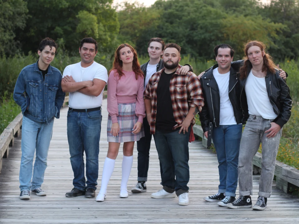 A group of seven people standing on a wooden path in a natural, wooded area. They are dressed casually, with varying styles including denim, plaid, and leather jackets.