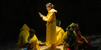 A woman in a yellow raincoat stands holding an object, surrounded by four people in yellow raincoats sitting or crouching on a dimly lit stage.