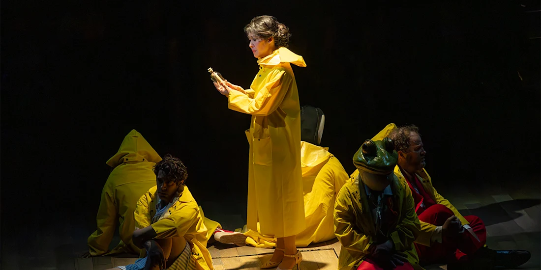 A woman in a yellow raincoat stands holding an object, surrounded by four people in yellow raincoats sitting or crouching on a dimly lit stage.