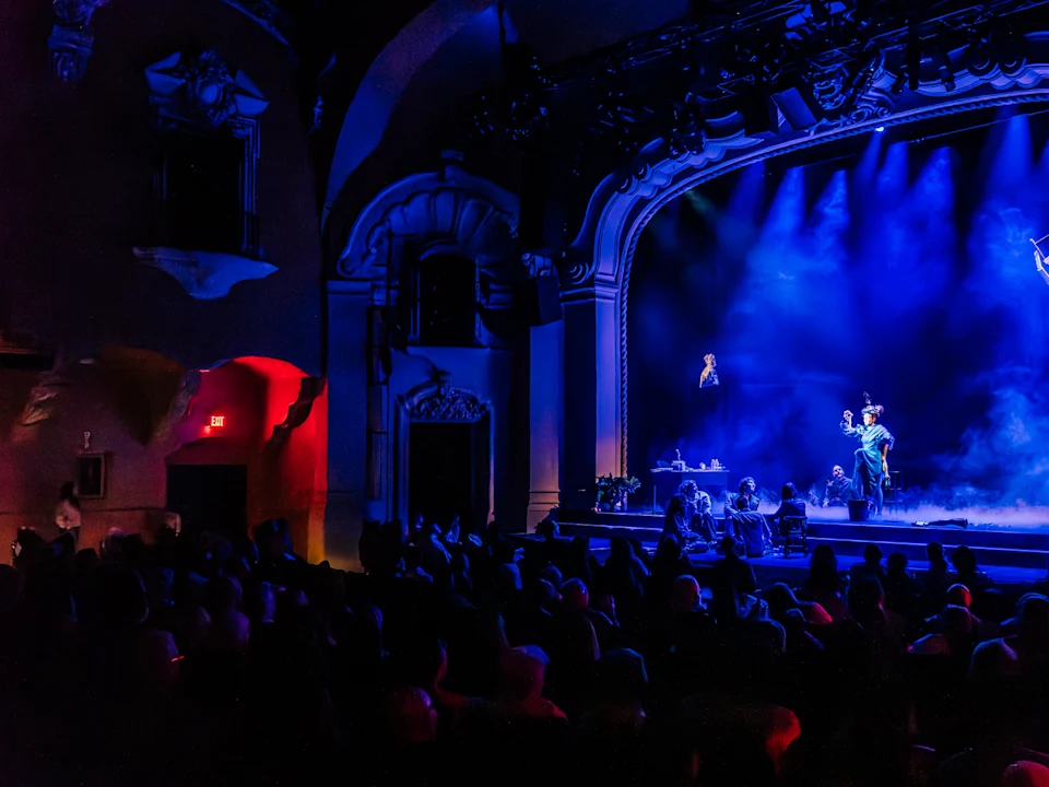 Production photo of ha ha ha ha ha ha ha, showing audience watches a theatrical performance on a stage lit with blue and purple lights, with actors in costume and atmospheric smoke effects.
