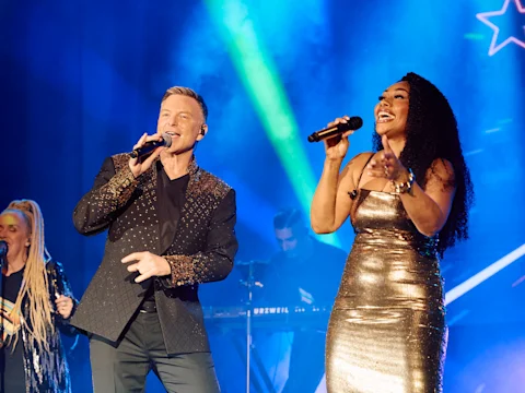 Tim Campbell (left) and Paulini (right) perform on stage with microphones, wearing stylish outfits. A live band and colorful stage lights are visible in the background.
