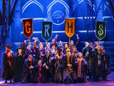 A group of actors in Hogwarts robes stand on stage beneath red, yellow, and green house banners, cheering with arms raised in front of a gothic-inspired set.