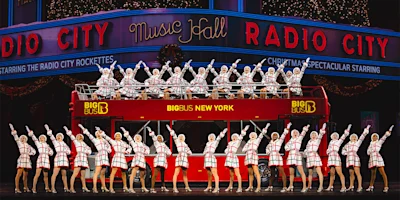 A large group of performers in matching red and white costumes pose in front of and on top of a double-decker bus on stage at Radio City Music Hall.