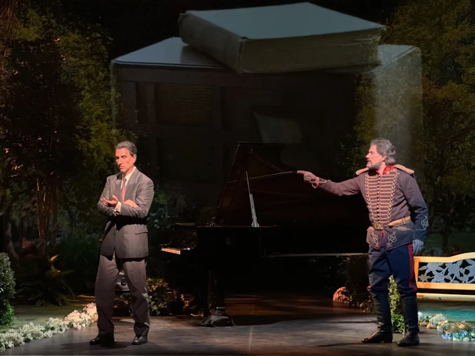 Two actors perform on stage; one in a suit gestures, while the other in a period costume points. A piano and garden scenery are in the background.