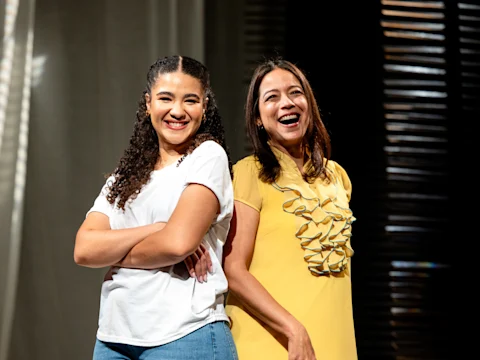 Two women stand back to back, smiling. One wears a white shirt and blue jeans with arms crossed, the other wears a yellow dress with ruffle details. Light and shadow appear in the background.