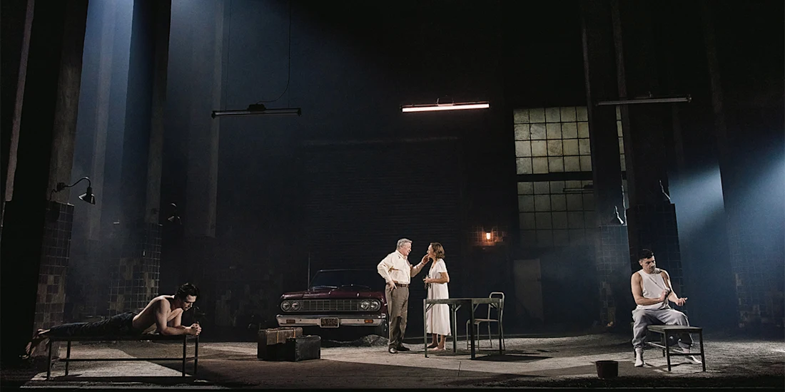 Four people are on a dimly lit stage with minimal props, including a red car, table, and chairs. Two people stand and talk while two others sit and lie on separate beds.