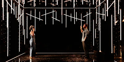Two people stand facing each other in a dark room illuminated by numerous vertical and horizontal light bars suspended from the ceiling.