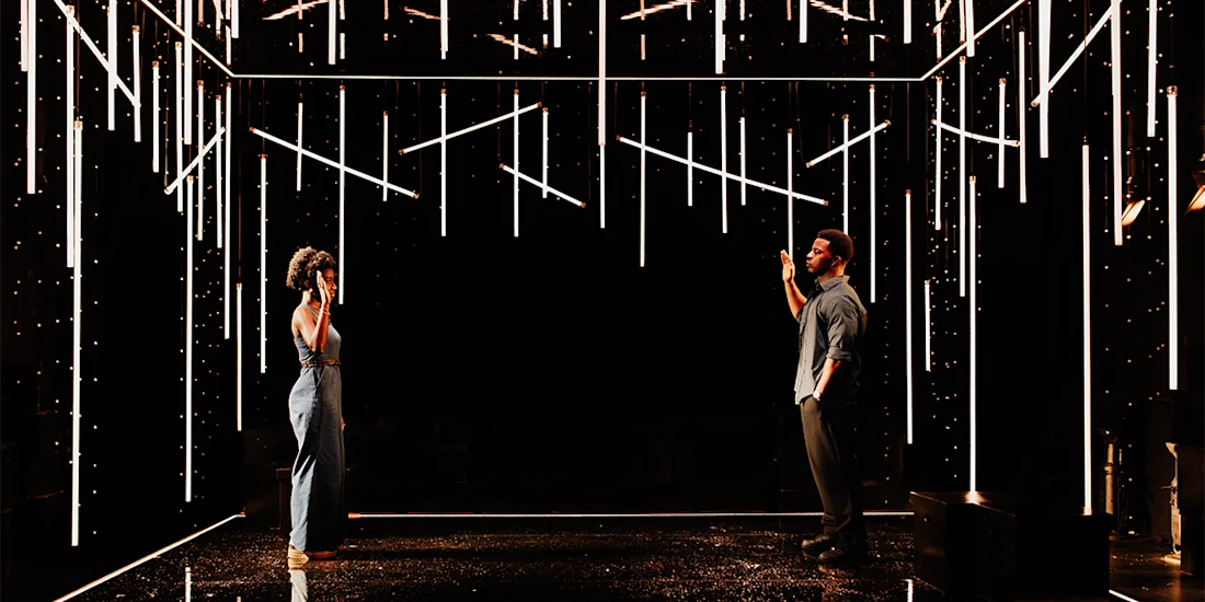 Two people stand facing each other in a dark room illuminated by numerous vertical and horizontal light bars suspended from the ceiling.