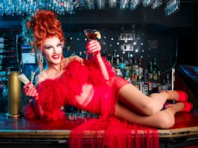 A person in a dramatic red outfit and wig is lying on a bar counter, holding a drink and a dollar bill, with bottles of alcohol in the background.