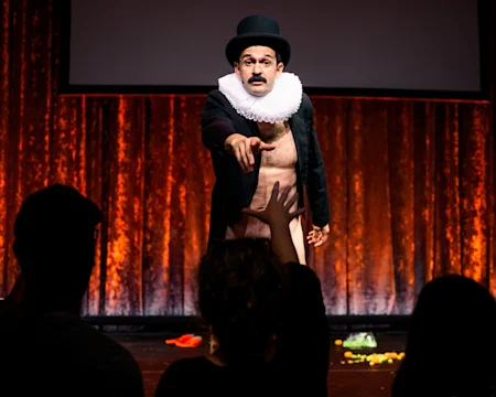 A performer in a top hat, ruff, and open coat, exposing his body, reaches toward an audience member on a stage with orange lighting and scattered props.