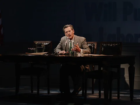 A man in a suit sits at a wooden table with papers, a microphone, and a water pitcher, speaking and gesturing with his hand under stage lighting.