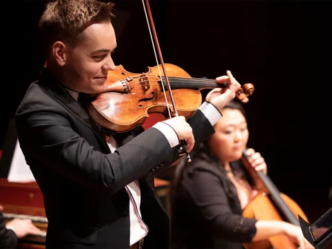 A violinist in a black suit plays the violin with focus, while a cellist in the background plays the cello.