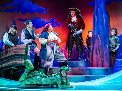 Cast members in pirate-themed costumes performing on a stage, including a person in a crocodile suit lying in the foreground.
