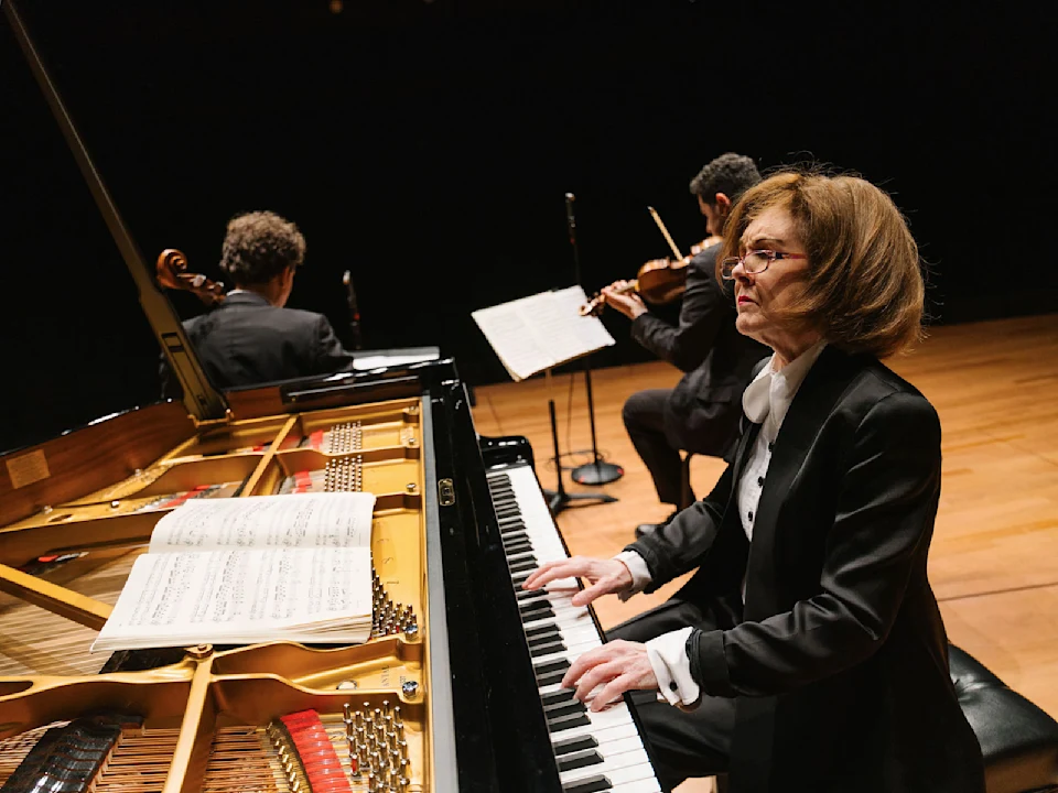 A woman in formal attire plays a grand piano with sheet music, accompanied by two musicians with string instruments on a stage.