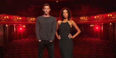Two people stand at the front of an empty theater with red seats and dim lighting, facing the camera.