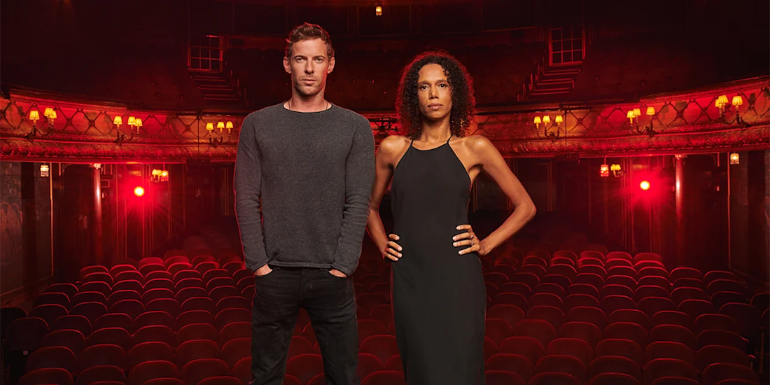 Two people stand at the front of an empty theater with red seats and dim lighting, facing the camera.