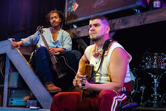 Two men sit on a stage with guitars, one on a staircase and the other in front, both looking intently ahead; stage lighting and musical instruments are visible in the background.