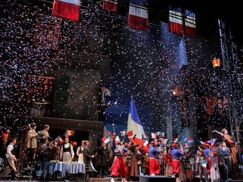 Performers on stage dressed in 19th-century attire wave French flags as red, white, and blue confetti falls from above, against a backdrop reminiscent of a historic French scene.