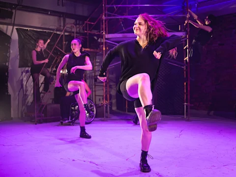 Two women in black outfits perform a high-energy dance routine on a lit stage, with two other people in the background near scaffolding.