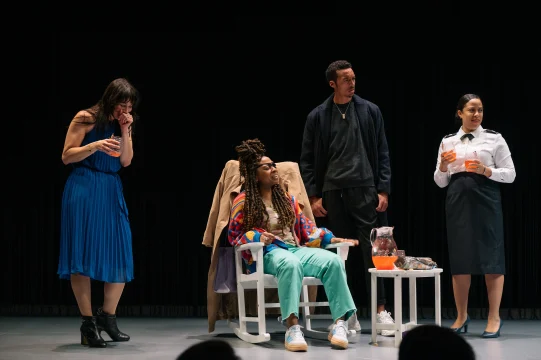 Four people are on stage, three women and one man. One woman in a blue dress is standing and laughing. Another woman with long braids is seated. The other man and woman are standing, holding drinks.