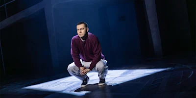 A person in a maroon sweatshirt and gray pants squats on a dimly lit stage, illuminated by a spotlight, with a dark, geometric background.