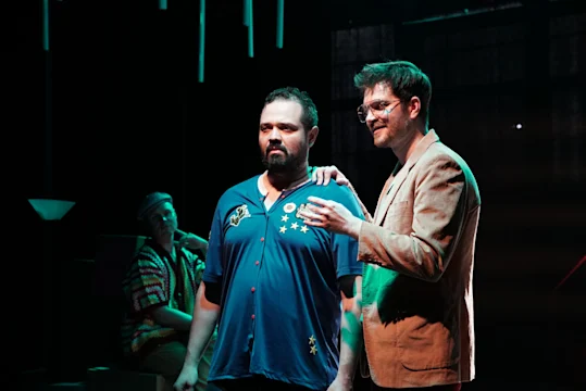Two men stand side by side on stage, one with a hand on the other's shoulder, while a third person sits in the background under dramatic lighting.