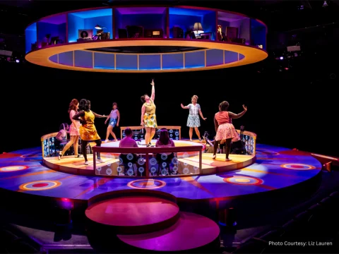 A brightly lit, colorful stage features several women in 1960s-style dresses dancing and singing. Circular platforms and vibrant hues dominate the modern set design.