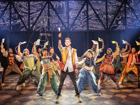 Production photo of & Juliet in Los Angeles, showing a group of costumed performers on stage, with one person in the center holding an arm up. They are surrounded by others, all holding papers. The backdrop features a collage of newspaper prints.