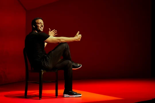 Production photo of Other in New York, showing a man in black clothes sits on a chair on a red stage, gesturing with both hands and smiling, with one leg crossed over the other.