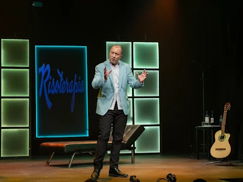A man in a light blue blazer stands on stage speaking, with a guitar, table, and chair nearby. The background displays illuminated panels and the word "Risoterapia.
